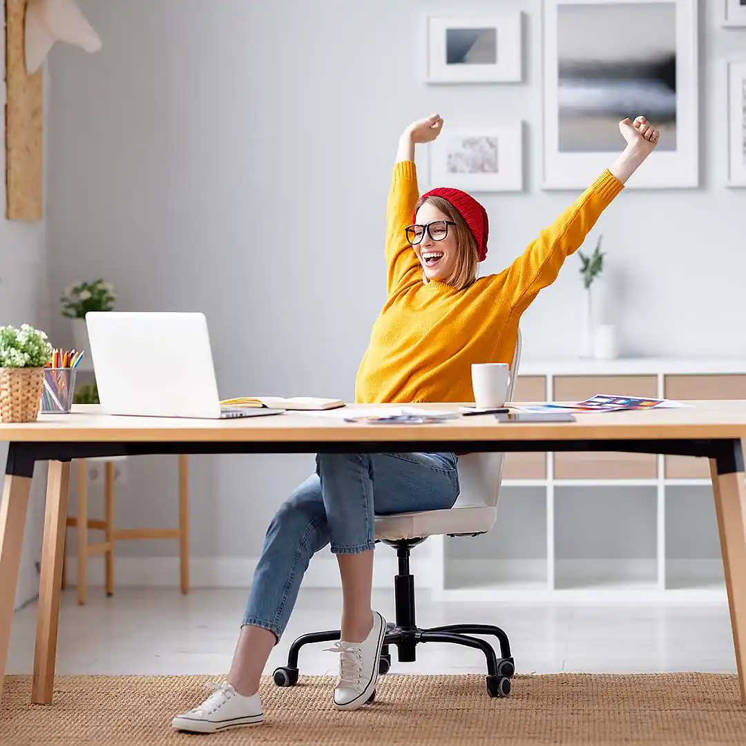 rubyni® Magnesium Junges Mädchen mit gelbem Pulli und roter Mütze und Brille sitzt zuhause am Schreibtisch vor ihrem Laptop und streckt sich fröhlich