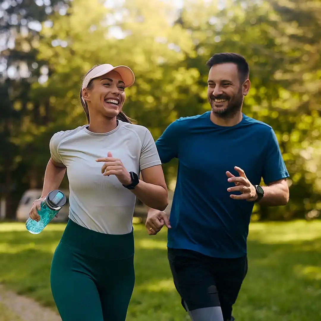 rubyni® Magnesium Eine sportliche junge Frau joggt mit einem sportlichen jungen Mann durch einen Park bei Sonnenschein. Die Frau hält eine Trinkflasche.