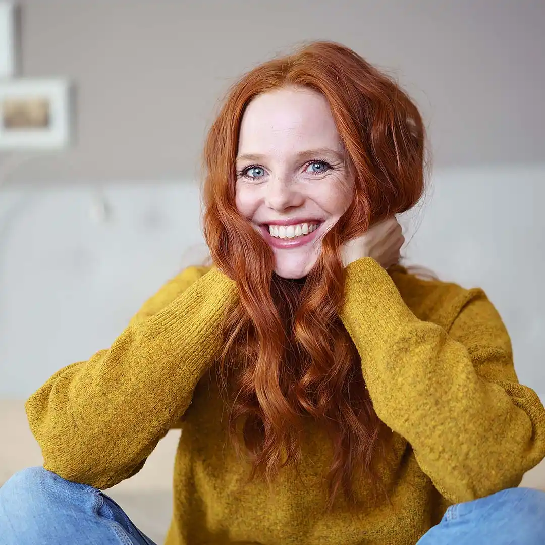rubyni® Selen – nicht nur für die Schilddrüse unverzichtbar Ein lachendes Mädchen mit langen roten Haaren und strahlend blauen Augen fasst sich an den Hals, sie trägt einen gelben Wollpulli und Jeans und sitzt in ihrem Wohnzimmer, das verschwommen zu erkennen ist.