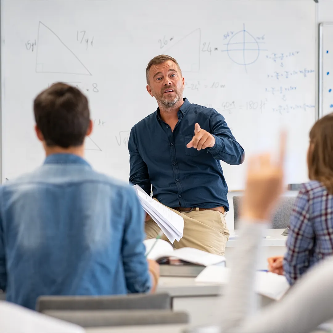 rubyni® Vitamin B KOMPLEX – für mehr Energie und bessere Nerven Man sieht ein Klassenzimmer, in dem ein Dozent vor Schülern unterrichtet. Er deutet mit dem Finger auf ein Mädchen, das die Hand hebt.