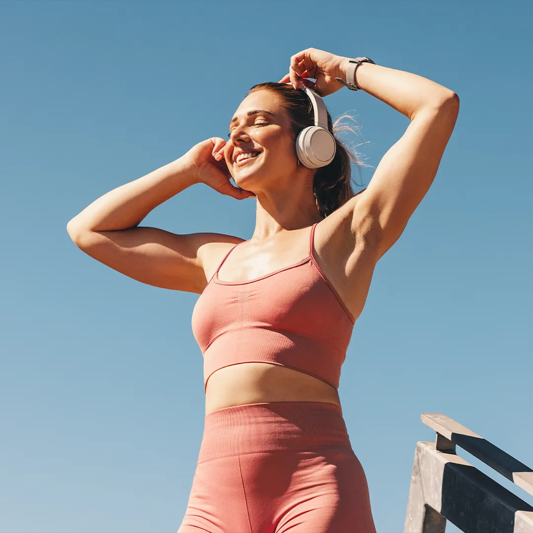 rubyni® Vitamin B KOMPLEX – für mehr Energie und bessere Nerven Eine junge Frau hört Musik aus weißen Kopfhörern, sie hebt die Hände hoch an die Kopfhörer und hat die Augen geschlossen. Sie genießt die Musik und ist sportlich mit einem lachsfarbenen hautengen Top und Hose bekleidet. Hinter ihr ist blauer Himmel.