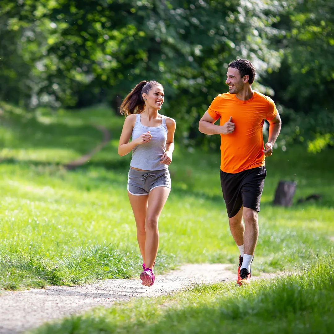 rubyni® Vitamin B KOMPLEX – für mehr Energie und bessere Nerven Eine junge Frau und ein junger Mann joggen auf einem Weg im Park. Sie lächeln sich zu. Er trägt ein oranges T-Shirt und eine kurze schwarze Hose, sie ein graues Top und eine graue kurze Hose. Sie hat ihre dunklen Haare zu einem Pferdeschwanz zusammengebunden. Man sieht den sandigen Weg, Wiese und Bäume. Die Sonne scheint.