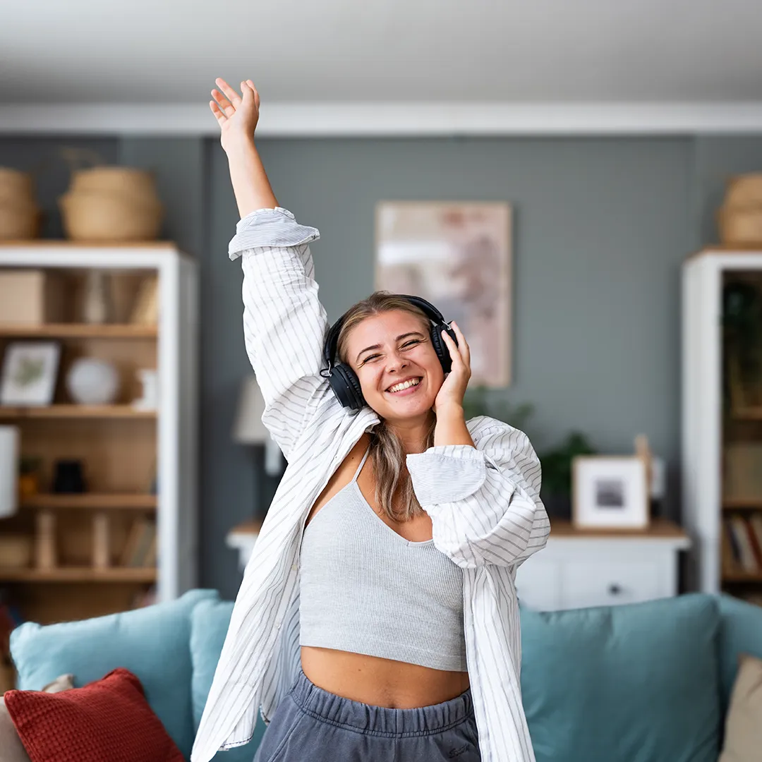 rubyni® Vitamin B12: Ihr Begleiter für mehr Energie im Alltag Ein glückliches Mädchen jubiliert in ihrem Wohnzimmer, sie streckt die Hand zur Decke, die andere Hand hält sie sich am Ohr, sie trägt schwarze Kopfhörer.