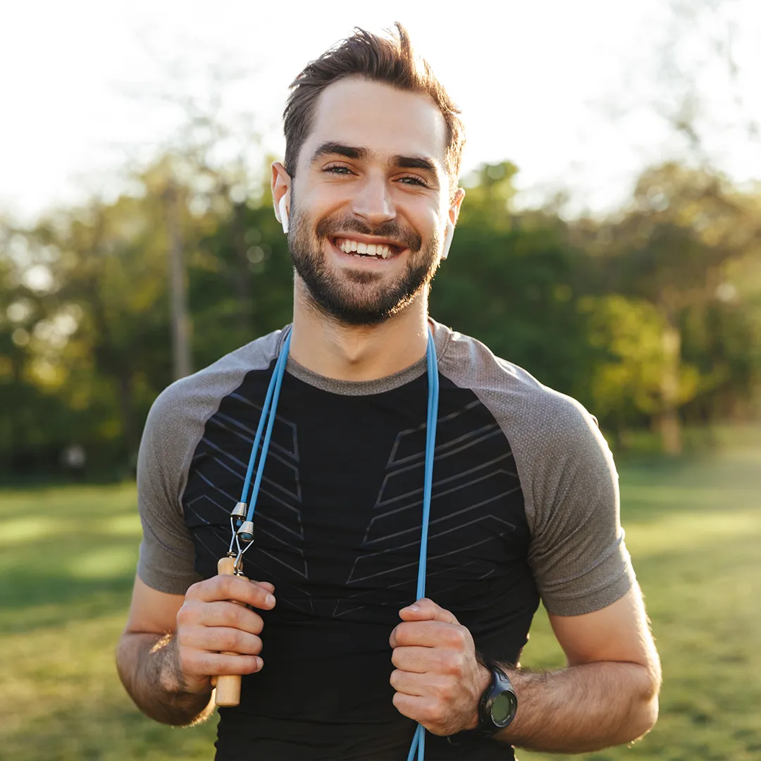 rubyni® Vitamin B12: Ihr Begleiter für mehr Energie im Alltag Ein junger Mann steht im Park, er ist sportlich angezogen, lächelt zufrieden und hält mit den Händen ein blaues Sprungseil, das er um den Hals hängen hat.