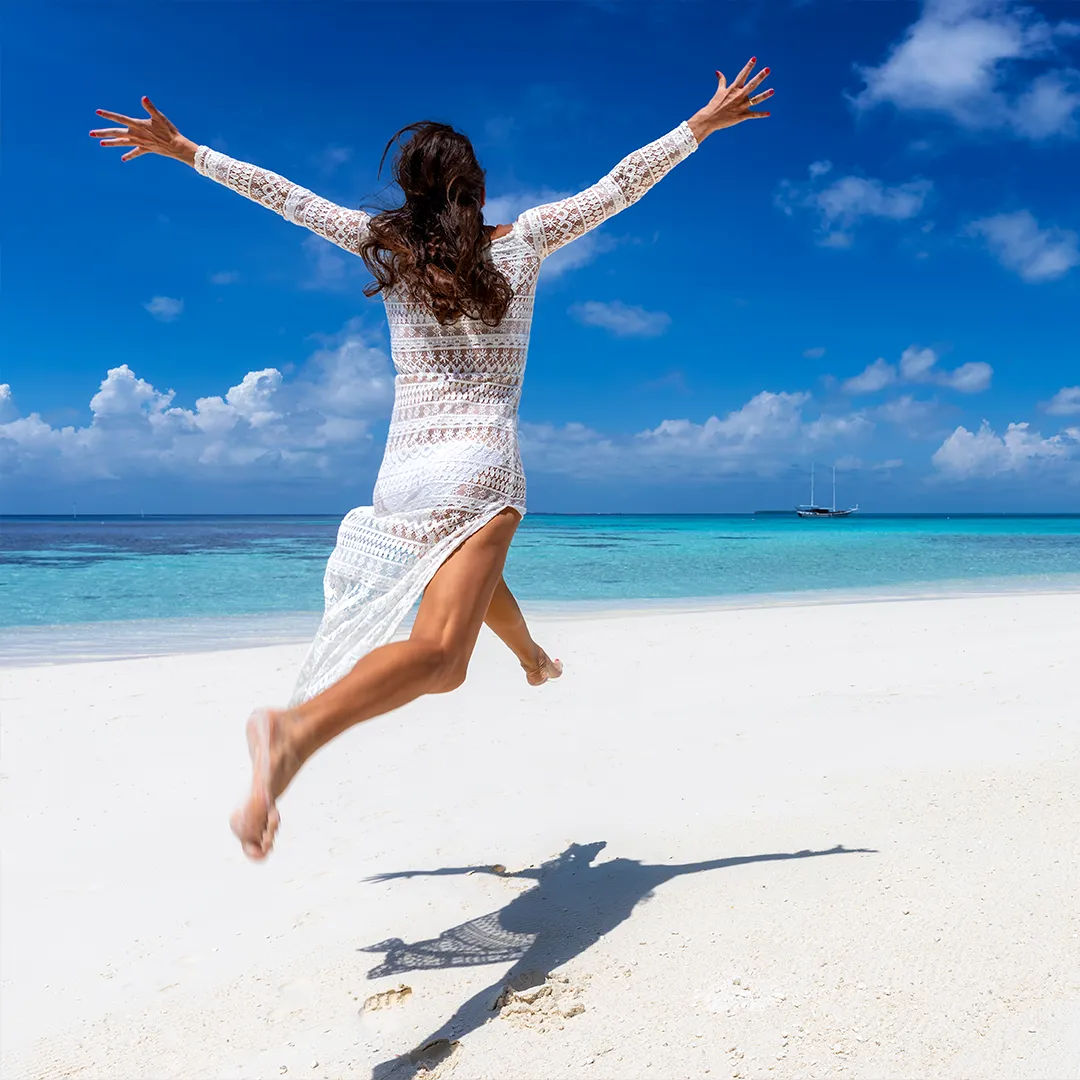 rubyni® Vitamin D – das Multitalent für eine gute Gesundheit Eine weiß gekleidete Frau im gehäkelten Sommerkleid springt am Meer auf strahlend weißem Sand. Sie macht einen Freudensprung. Dahinter sieht man das türkisblaue Meer.