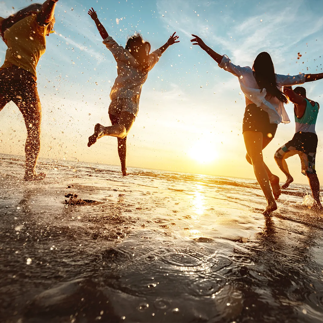 rubyni® Vitamin D – das Multitalent für eine gute Gesundheit Eine Gruppe von vier jungen Leuten, drei Mädchen, ein Junge, läuft jubelnd mit Armen nach oben am Strand durchs flache Meer, Wasser spritzt. Es ist Sonnenuntergang.