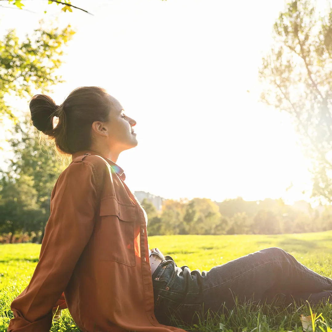 rubyni® Vitamin K: Unterstützung für starke Knochen Eine junge Frau sitzt auf einer Wiese im Park. Sie hat die Augen geschlossen und neigt den Kopf nach hinten, sie stützt sich mit den Armen hinter dem Rücken auf. Sie ist barfuß und trägt ein oranges offenes Hemd über ihrer Kleidung und einer Jeans. Die Sonne scheint und sie macht einen sehr entspannten Eindruck.
