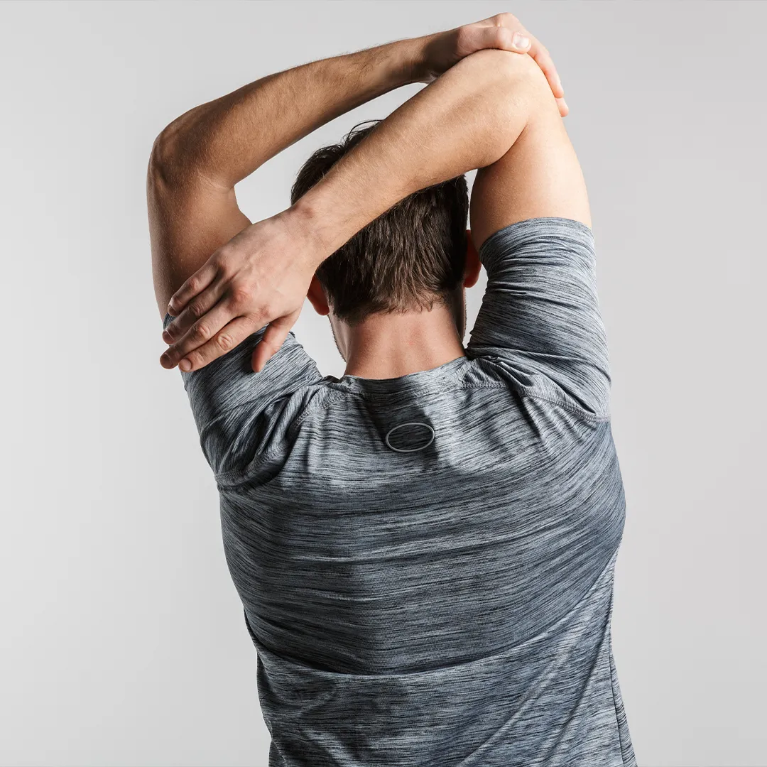 rubyni® Vitamin K: Unterstützung für starke Knochen Ein junger sportlicher Mann dehnt seine Schultermuskeln. Er reckt dazu seine Arme nach oben und zieht mit der linken Hand am rechten Oberarm. Er trägt ein graues Sport-Shirt.