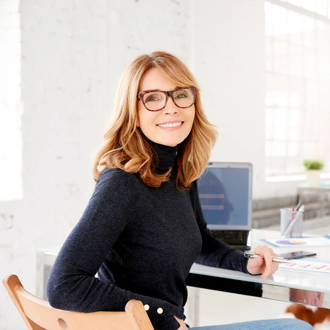 rubyni® Zink – das Spurenelement für Gesundheit und Schönheit Eine Business Woman / Geschäftsfrau dreht sich an ihrem Schreibtisch zur Kamera um und lächelt souverän mit einem Stift in der Hand in die Kamera. Sie trägt eine Brille und offenes blondes halblanges sowie einen hochgeschlossenen dunkelblauen Pulli.