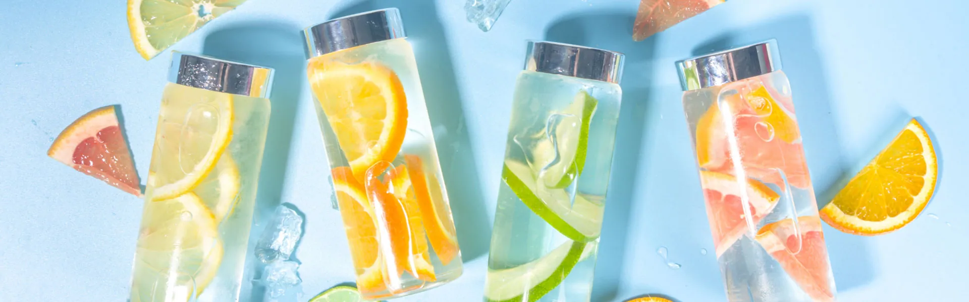 Durchsichtige Glas-Trinkflaschen liegen auf einer blauen Tischplatte, Sicht von oben auf die Flaschen, silberner Aludeckel. In den Flaschen befindet sich Wasser mit Zitronen-, Limetten- und Orangenschnitten. Es liegen auch noch ein paar Eiswürfel auf dem Tisch.