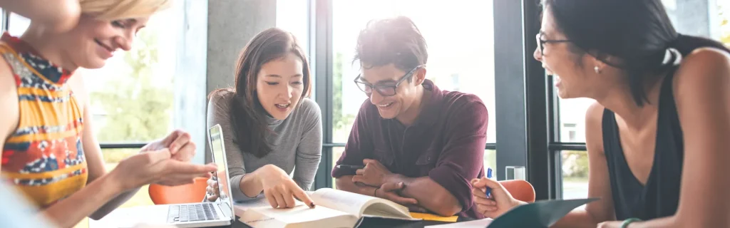 Junge Leute sitzen an einem Tisch und arbeiten, sie lachen dabei und sind gut gelaunt. Auf
dem Tisch sieht man ein Laptop und ein Buch, auf das ein Mädchen deutet.