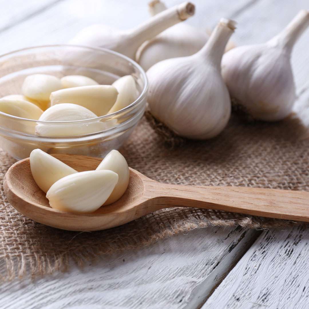 Auf einem weißen Holztisch liegen auf einem Stück Jute vier Knoblauch, in einem Glasschälchen liegen geschälte Knoblauchzehen. Auf einem Holzlöffel liegen drei geschälte Knoblauchzehen. 