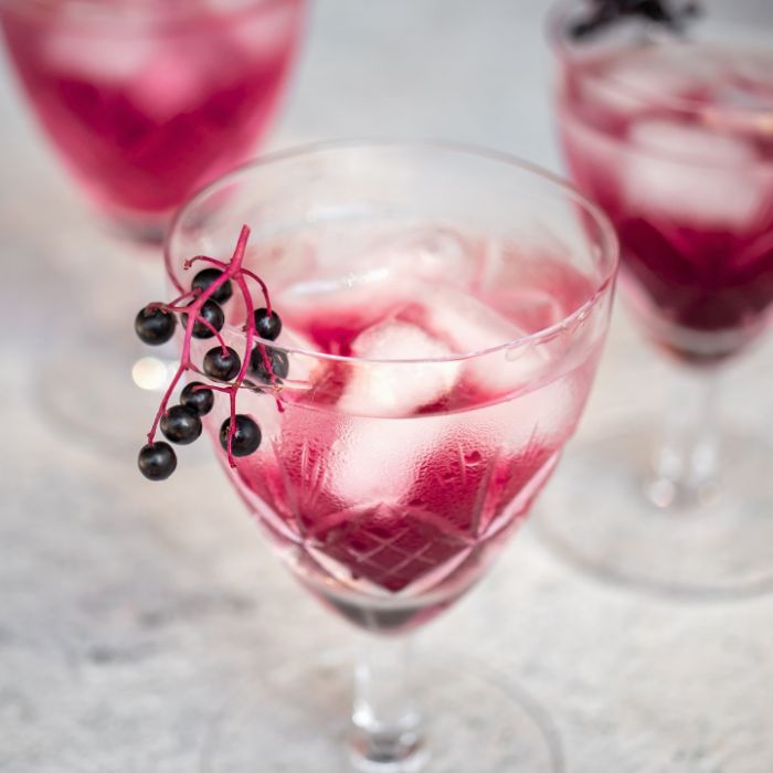 Ein abgeschnittenes Glas, das man von oben sieht. Darin befinden sich Eiswürfel und eine rote Flüssigkeit, am Rand des Glases hängt ein Büschel Holunderbeeren als Deko.