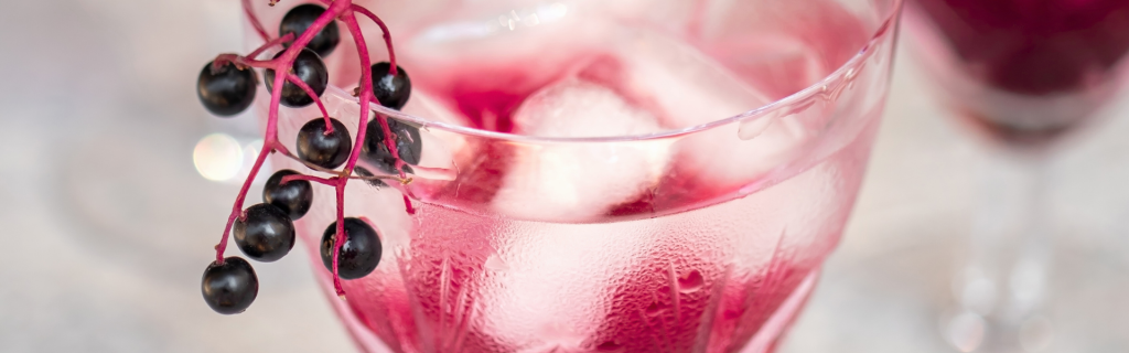 Ein abgeschnittenes Glas, das man von oben sieht. Darin befinden sich Eiswürfel und eine rote Flüssigkeit, am Rand des Glases hängt ein Büschel Holunderbeeren als Deko.