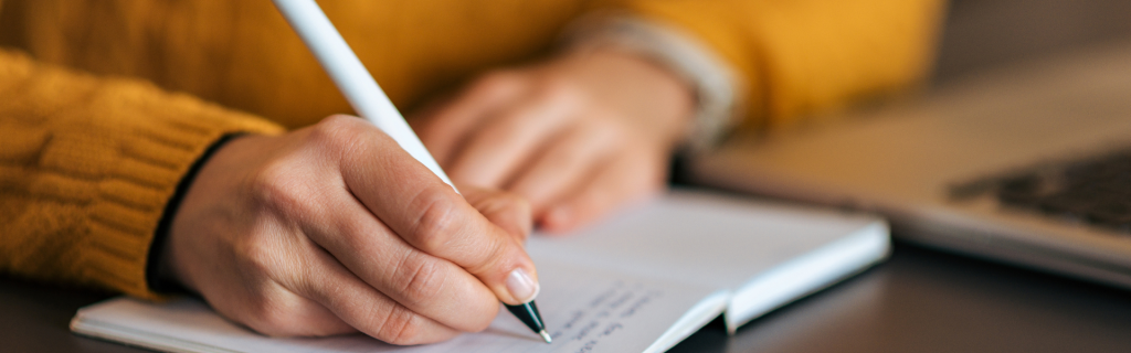 Man sieht ausschnittsweise eine Hand, die einen weißen Stift hält und in ein Kalenderbuch schreibt. Man sieht auch noch ausschnittsweise, dass diese Person einen gelben Pulli trägt.