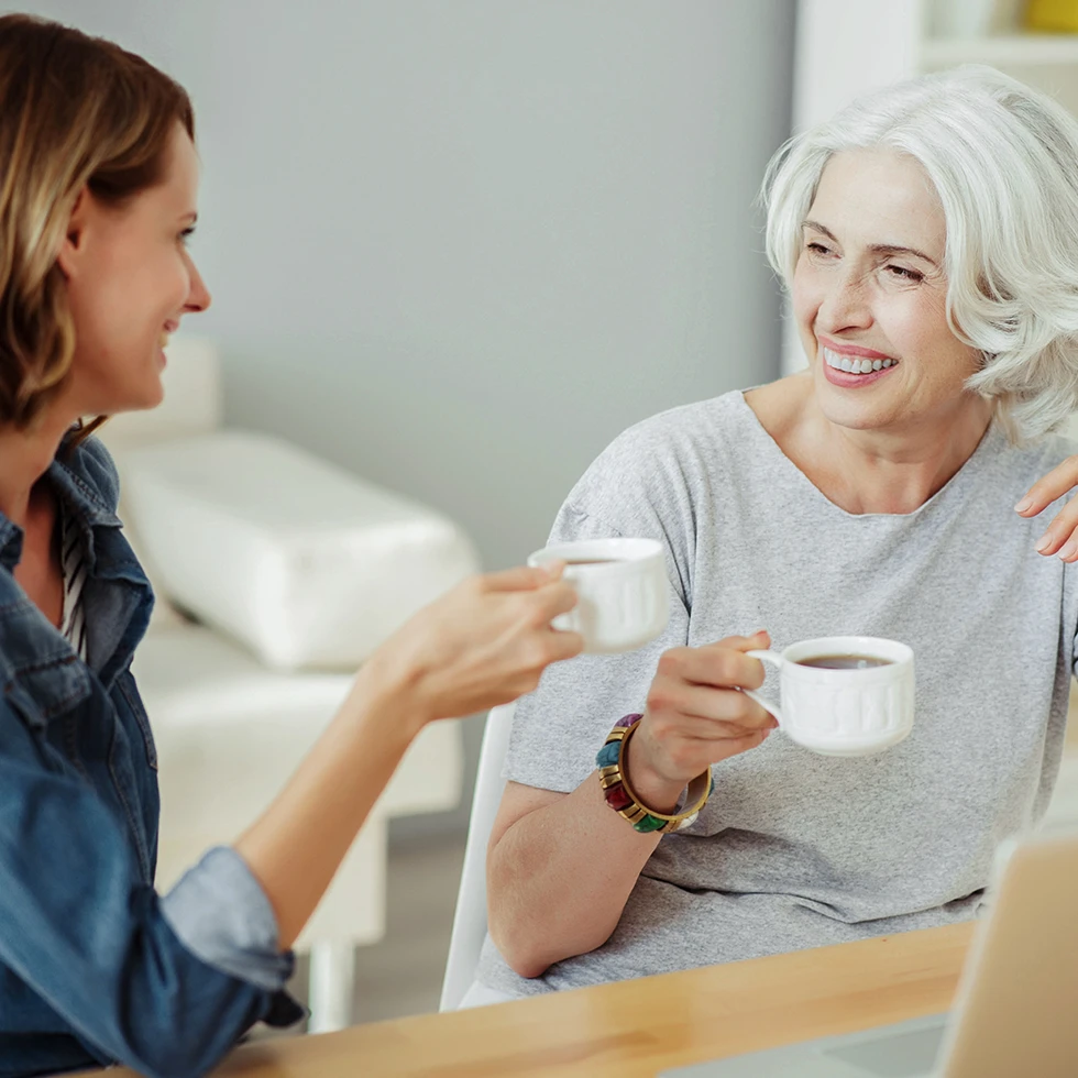 Eine jüngere und eine ältere Frau sitzen gemeinsam an einem Tisch, trinken Kaffee und unterhalten sich lächelnd. Vor ihnen steht ein Laptop, die Szene vermittelt Austausch, Vertrauen und generationsübergreifende Nähe.