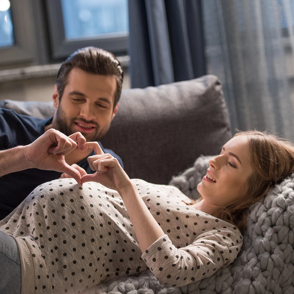 Ein werdendes Elternpaar entspannt gemeinsam auf einem Sofa. Die schwangere Frau liegt gemütlich angelehnt, während der Mann liebevoll ein Herz mit den Händen über ihrem Babybauch formt. Die Szene vermittelt Nähe, Vorfreude und familiäre Geborgenheit.