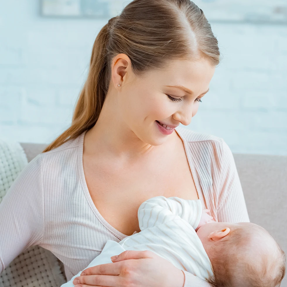 Eine junge Mutter sitzt entspannt auf einem Sofa und stillt ihr Baby. Sie blickt liebevoll auf ihr Kind herab, das in eine helle Decke gehüllt ist. Die helle, ruhige Umgebung unterstreicht die intime und fürsorgliche Atmosphäre.