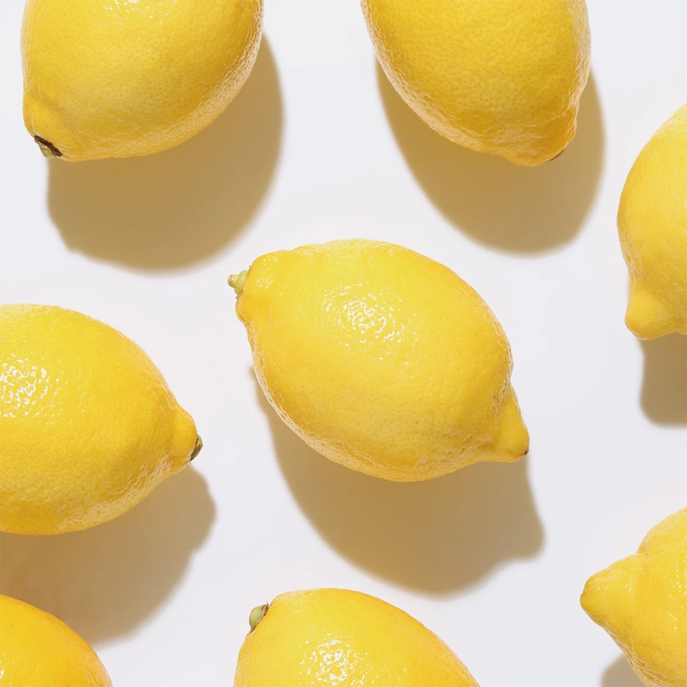 Mehrere frische, gelbe Zitronen sind gleichmäßig auf hellem Hintergrund angeordnet. Die Früchte wirken glänzend und saftig, mit deutlich sichtbarer, strukturierter Schale und weichen Schatten, die ein frisches, natürliches Erscheinungsbild unterstreichen.