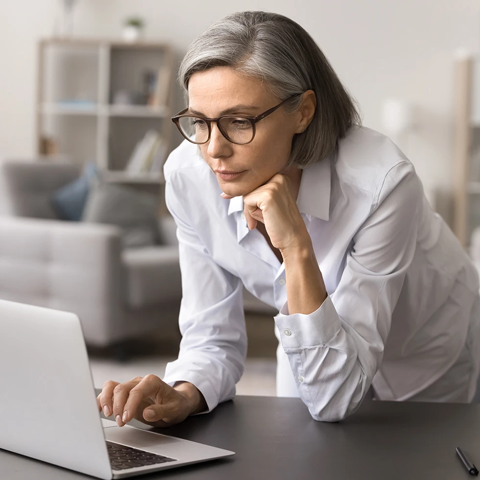 Eine Frau mit grauem Haar und Brille lehnt sich nachdenklich über einen Laptop. Sie wirkt konzentriert und fokussiert in einer modernen, hellen Wohnumgebung.