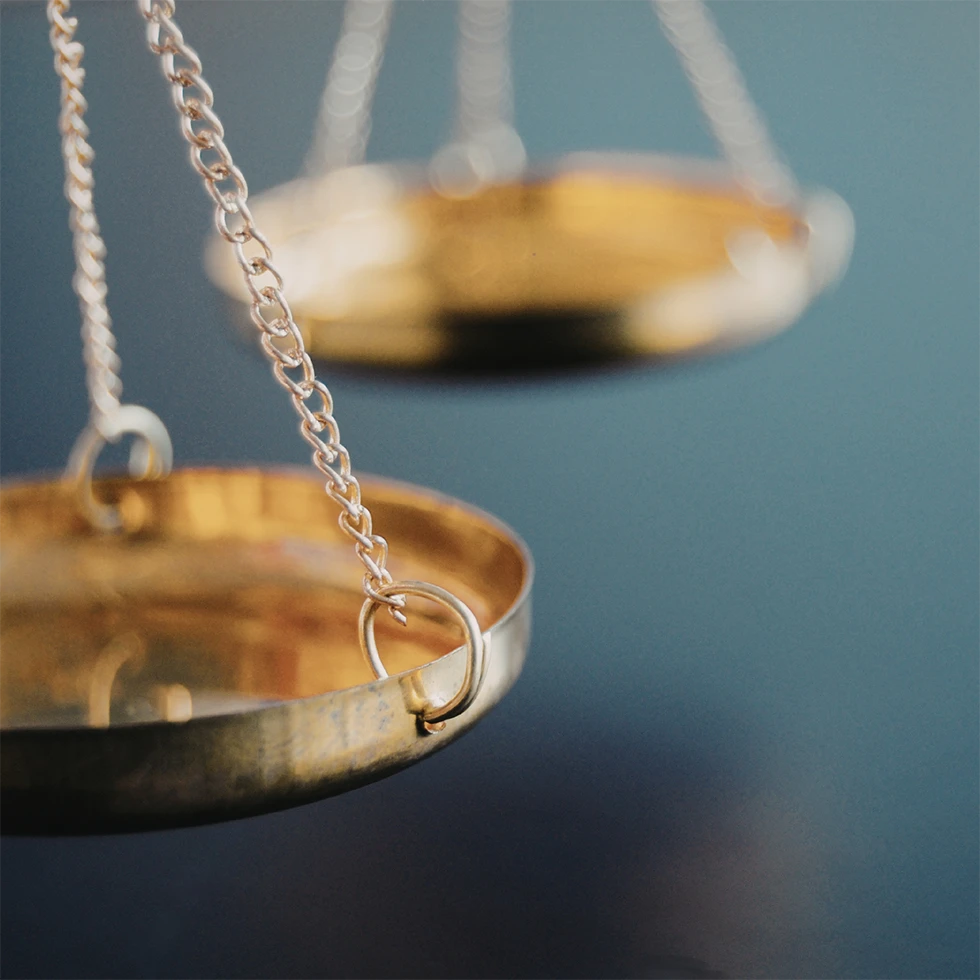 Nahaufnahme einer goldfarbenen Waage mit filigranen Ketten vor ruhigem, blauem Hintergrund. Eine der Waagschalen ist im Fokus, während die zweite unscharf im Hintergrund erscheint. Das Motiv symbolisiert Gleichgewicht und Balance.