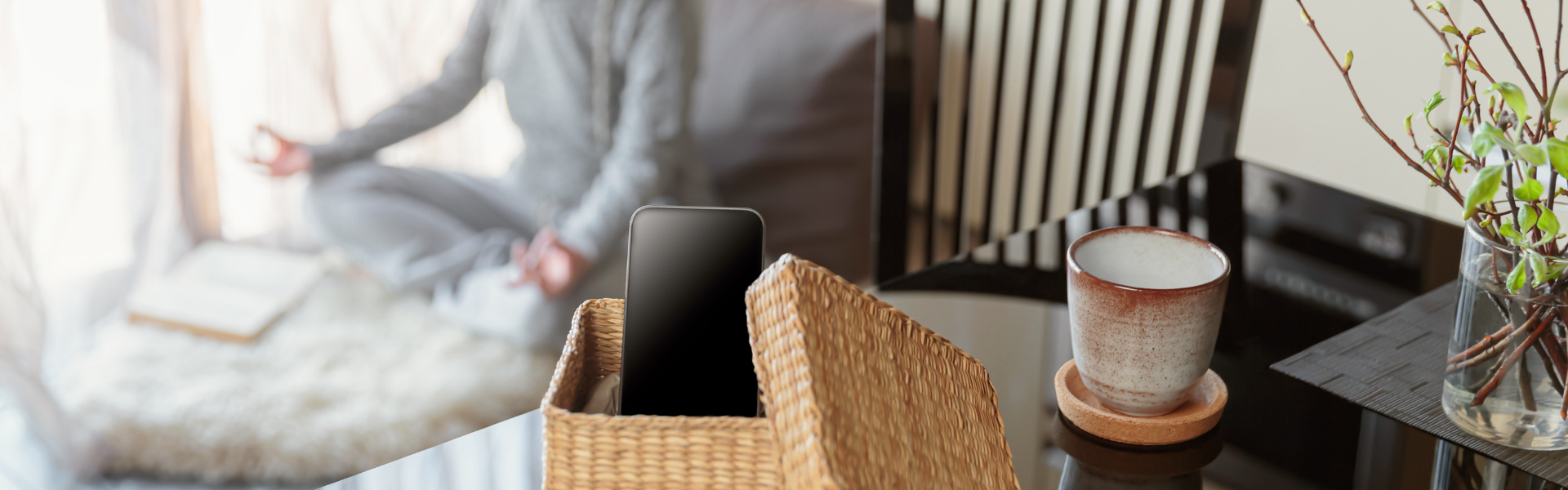 Headerbild für den rubyni Blog-Beitrag „Digital Detox“ ein Smartphone liegt in einem geflochtenen Weidenkörbchen auf einem schwarzen Glastisch. Daneben steht eine Tasse auf einem Untersetzer. Man sieht auch noch eine Vase mit einigen Zweigen. Im Hintergrund meditiert eine Frau in einem grauen Hausanzug auf einem Schaffell, man sieht sie nur auszugsweise und verschwommen.