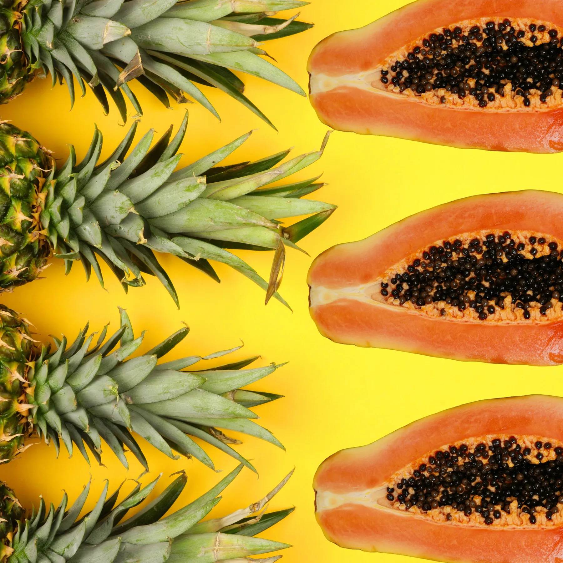 Mehrere halbierte Papayas mit sichtbaren schwarzen Kernen liegen auf einem gelben Hintergrund. Daneben sind die grünen Blattkronen von Ananasfrüchten angeordnet. Die farbenfrohe Kombination aus Orange, Grün und Gelb vermittelt Frische, Exotik und natürliche Fruchtzutaten.