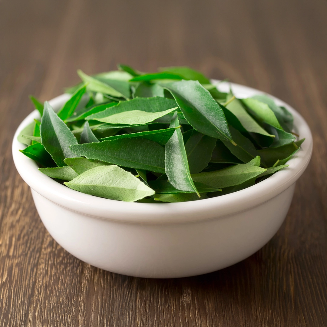 Eine Schale mit frischen, sattgrünen Curryblättern auf einer Holzoberfläche. Die Blätter wirken aromatisch und naturbelassen und stehen sinnbildlich für hochwertige pflanzliche Rohstoffe sowie deren ursprüngliche Qualität.