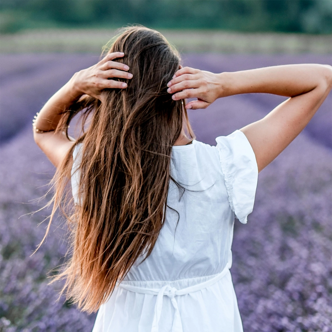 Eine Frau steht in einem weitläufigen Lavendelfeld und fährt sich mit beiden Händen durch ihr langes, gesund wirkendes Haar, das im natürlichen Licht weich glänzt und für Vitalität sowie Pflege von innen heraus steht.