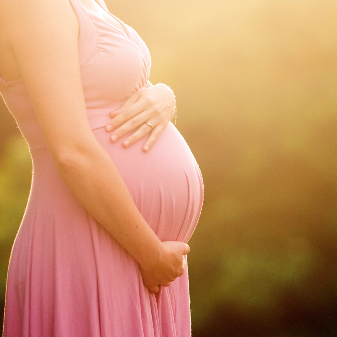 Eine schwangere Frau hält liebevoll ihren Bauch, während warmes Sonnenlicht die Szene durchflutet und ein Gefühl von Fürsorge, natürlicher Entwicklung und innerer Balance vermittelt.