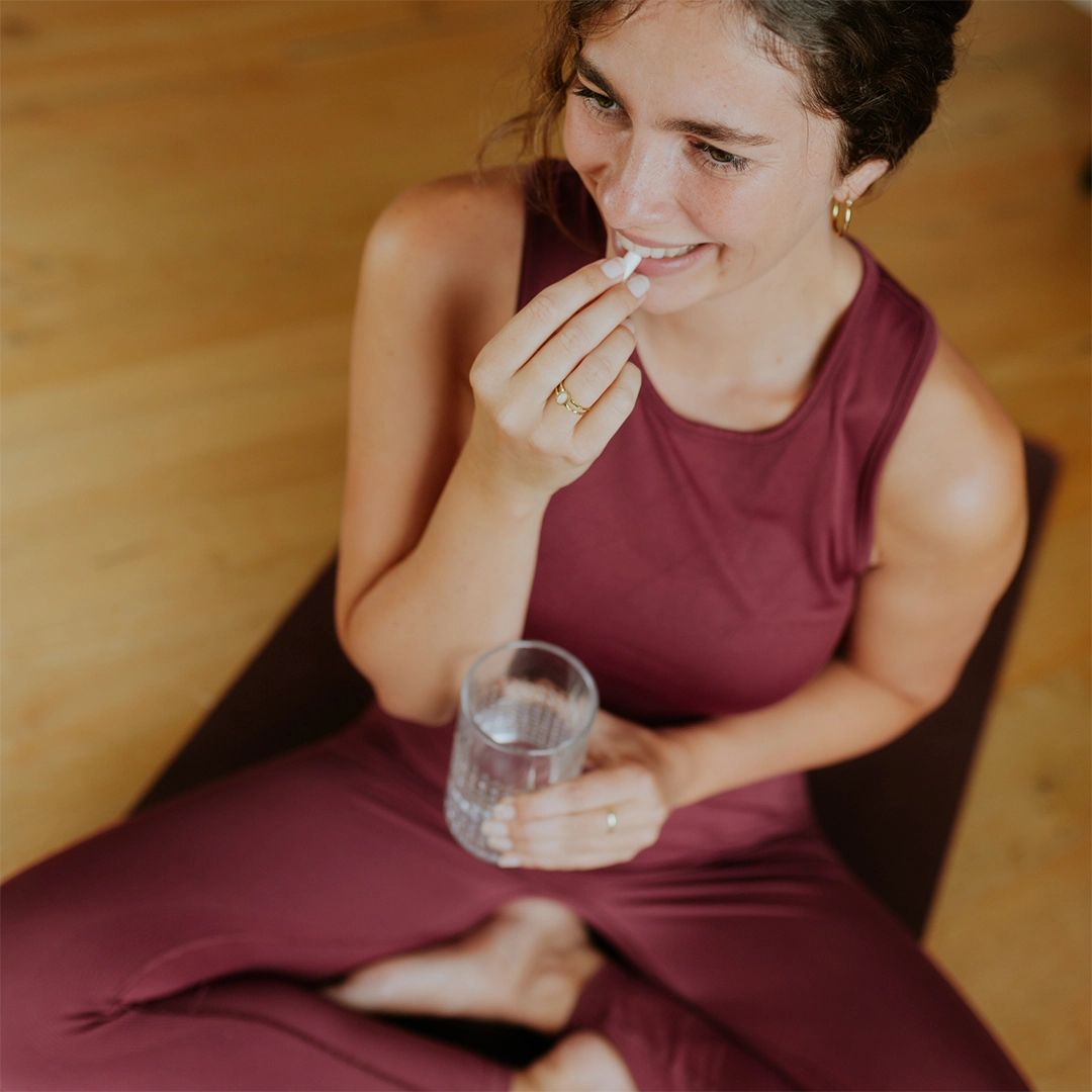 Eine junge Frau sitzt entspannt im Schneidersitz und nimmt eine Kapsel ein, während sie ein Glas Wasser in der Hand hält. Die Szene vermittelt eine bewusste, ruhige Einnahmeroutine und steht für die einfache Integration von Nahrungsergänzung in den Alltag.