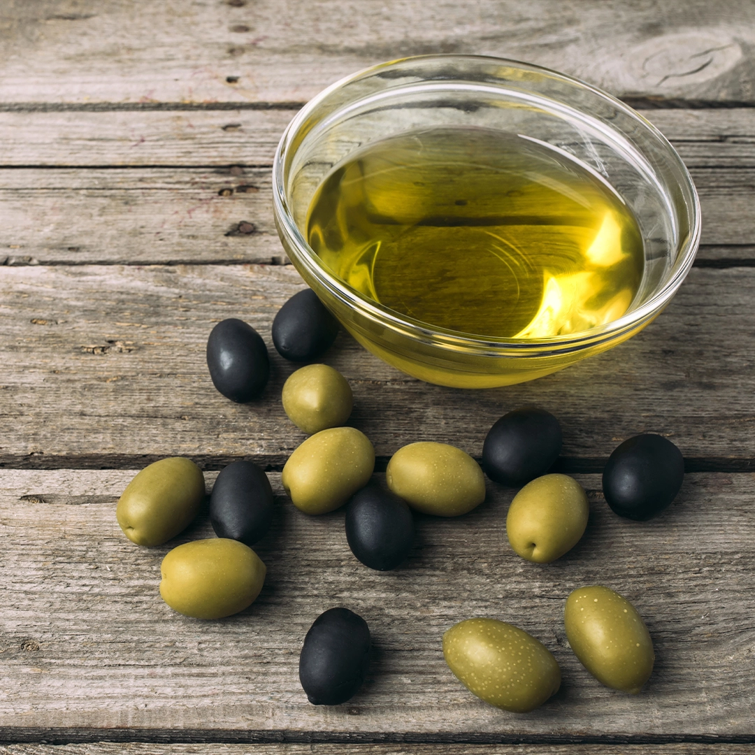 Eine Glasschale mit goldenem Olivenöl steht auf einer rustikalen Holzoberfläche, umgeben von grünen und schwarzen Oliven. Das Bild vermittelt Natürlichkeit und verweist auf hochwertige pflanzliche Inhaltsstoffe sowie traditionelle Rohstoffe.