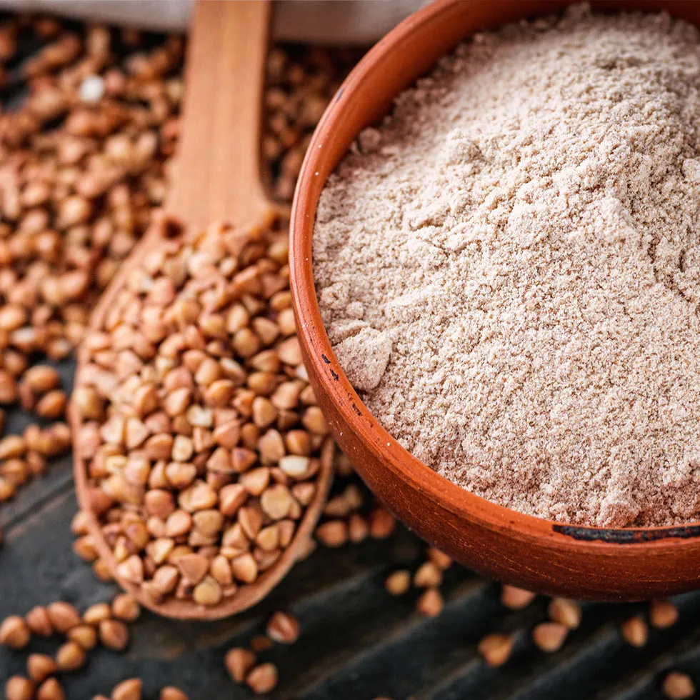 Eine Schale mit feinem Buchweizenkeimpulver steht auf einem rustikalen Holztisch. Daneben liegen ganze Buchweizenkörner und ein Holzlöffel, der mit den Körnern gefüllt ist. Die Darstellung betont natürliche, pflanzliche Rohstoffe und deren ursprüngliche Form.