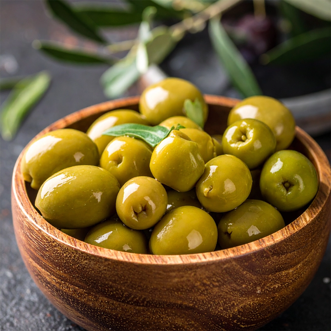 Eine Schale mit glänzenden grünen Oliven wird von frischen Olivenzweigen begleitet und steht für natürliche pflanzliche Inhaltsstoffe sowie wertvolle Nährstoffe aus mediterranen Quellen.