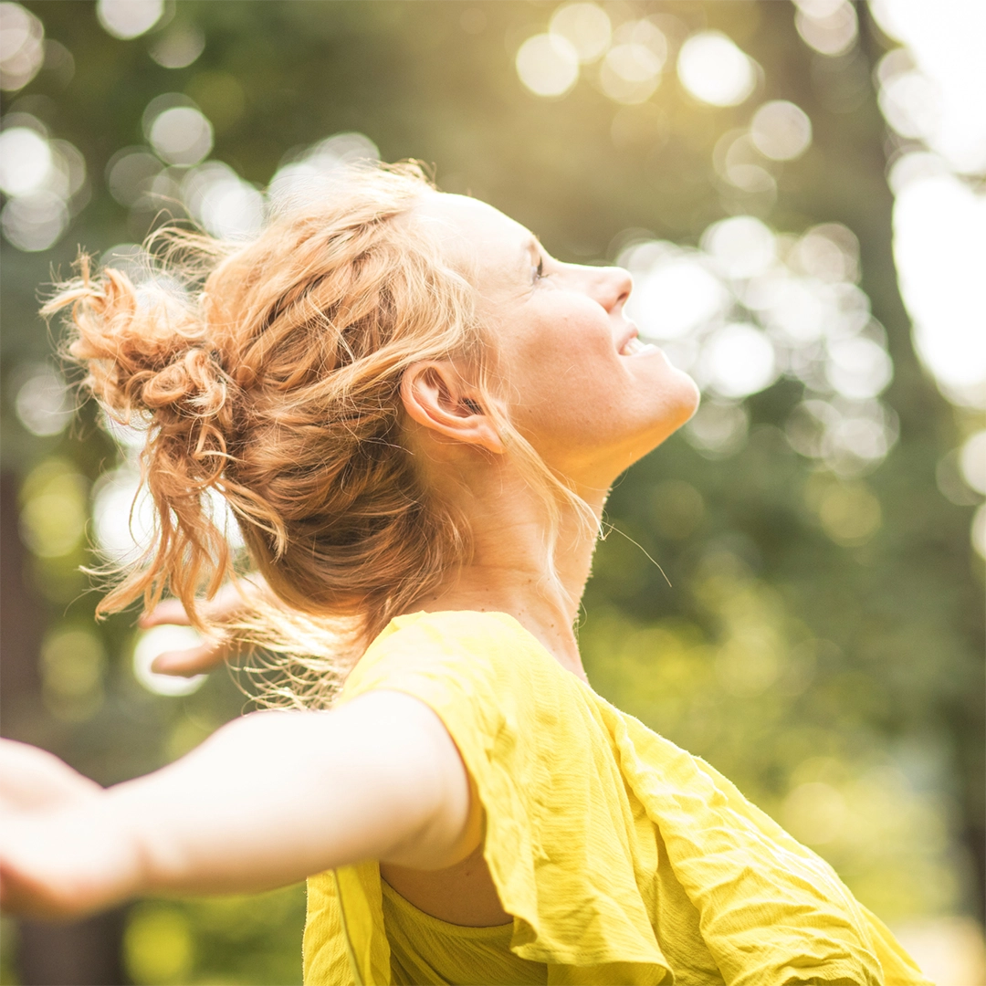 Eine Frau steht im Freien mit geschlossenen Augen und ausgebreiteten Armen im warmen Sonnenlicht. Die Szene vermittelt ein Gefühl von Energie, Leichtigkeit und innerer Balance und symbolisiert Wohlbefinden sowie neue Vitalität.