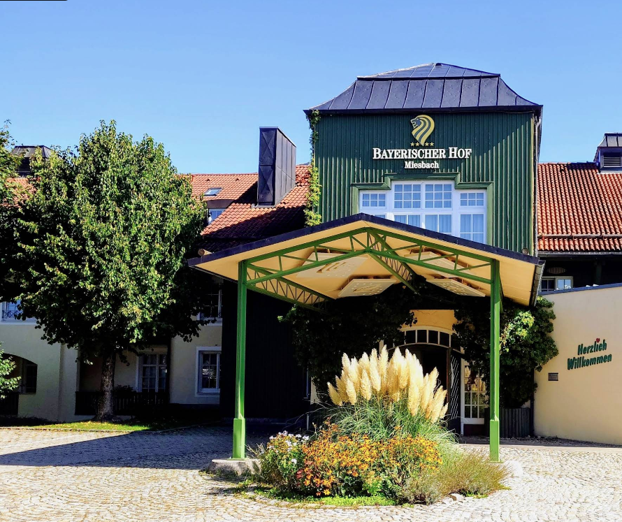 Außenansicht des Bayerischen Hofs in Miesbach an einem sonnigen Tag. Das Gebäude mit grünem Turm trägt den Schriftzug „Bayerischer Hof Miesbach“. Vor dem Eingang befindet sich ein überdachter Eingangsbereich mit grünen Stützen, davor ein bepflanztes Beet und ein gepflasterter Vorplatz. Rechts am Gebäude steht „Herzlich Willkommen“.