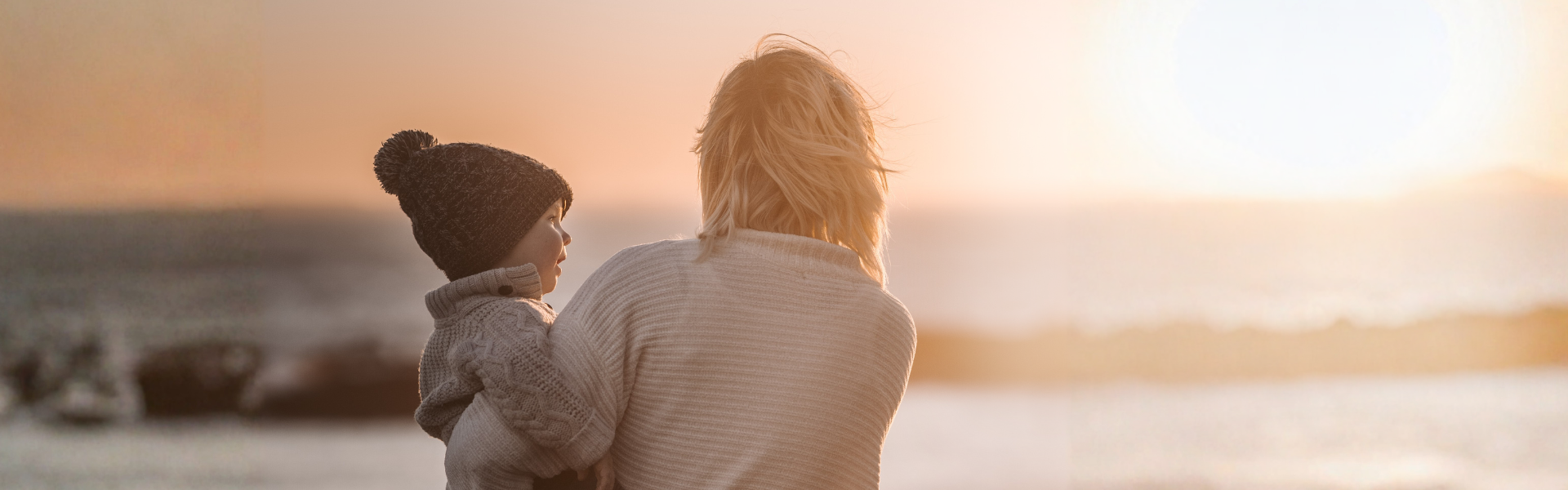 Titelbild zur Illustration des Blogbeitrags „Angeborenes und erworbenes Immunsystem Eine Mutter hält ihr kleines Kind auf dem Arm und blickt mit ihm gemeinsam auf das Meer. Die Szene ist in warmes Abendlicht getaucht, was eine ruhige, geborgene und familiäre Atmosphäre vermittelt.