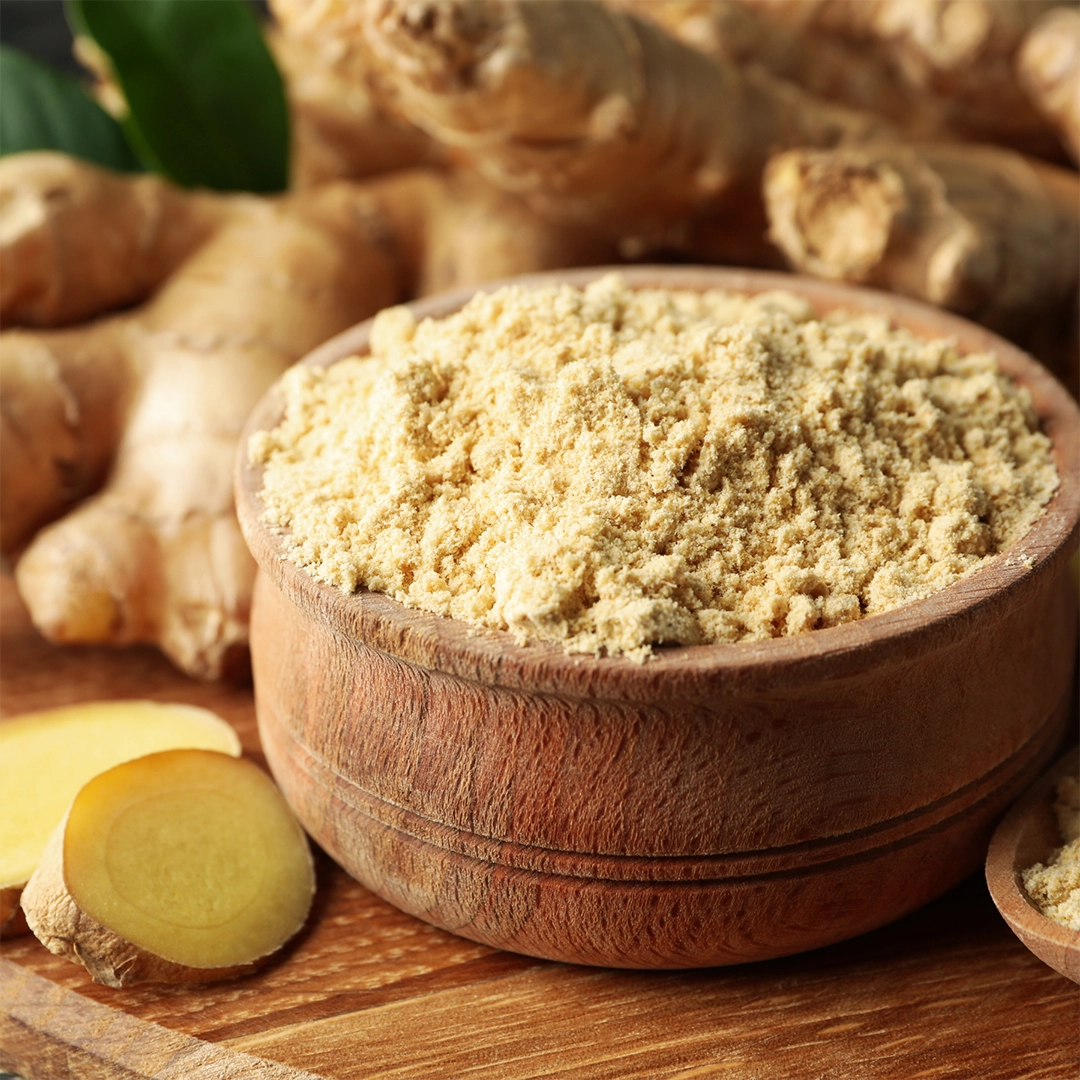 Fein gemahlenes Ingwerpulver in einer runden Holzschale, umgeben von frischen Ingwerwurzeln und einzelnen Ingwerscheiben auf einer Holzoberfläche.