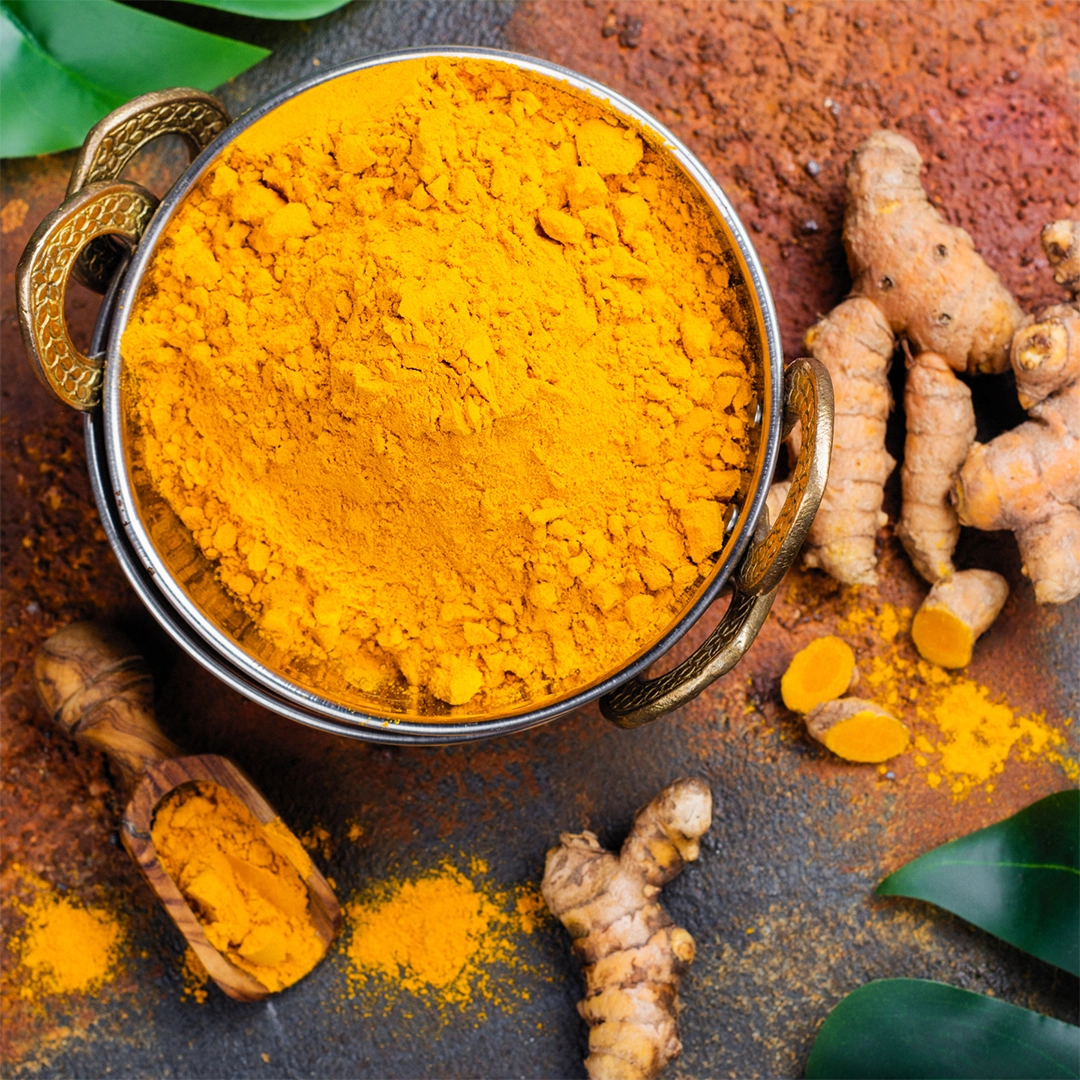 Leuchtend gelbes Curcumapulver in einer Metallschale, umgeben von frischen Curcuma-Knollen, etwas verstreutem Pulver und grünen Blättern auf dunkler Oberfläche.