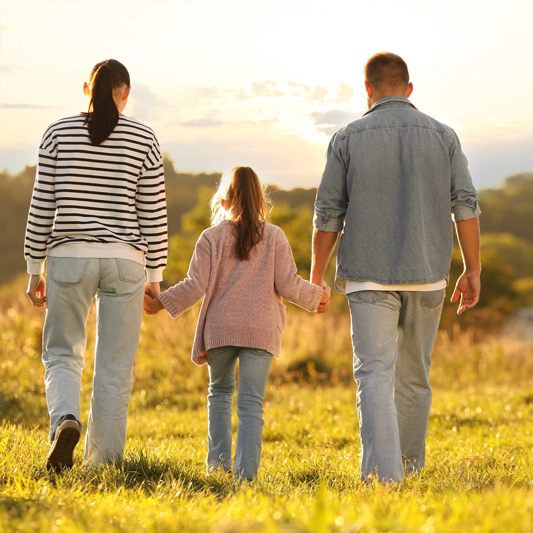 Familie mit Kind geht Hand in Hand über eine Wiese im warmen Sonnenlicht und genießt gemeinsam einen Spaziergang in der Natur.