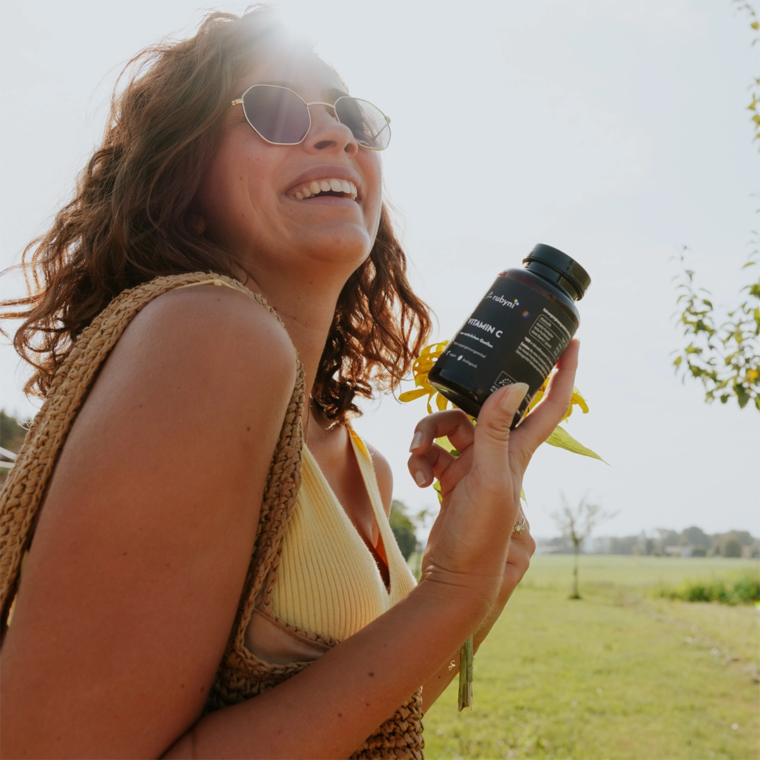 Frau mit Sonnenbrille lächelt im Sonnenlicht und hält eine PET-Dose rubyni Vitamin-C-Nahrungsergänzungsmittel in der Hand, während sie draußen in der Natur steht.