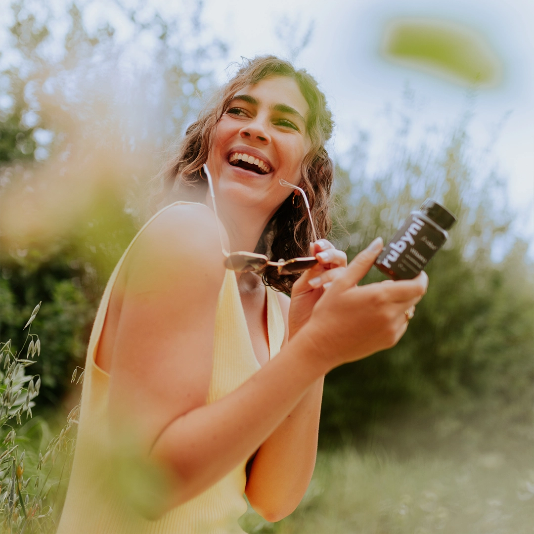 Frau sitzt lachend im Grünen, hält eine PET-Dose rubyni Vitamin D in der Hand und genießt die Natur bei Sonnenschein.