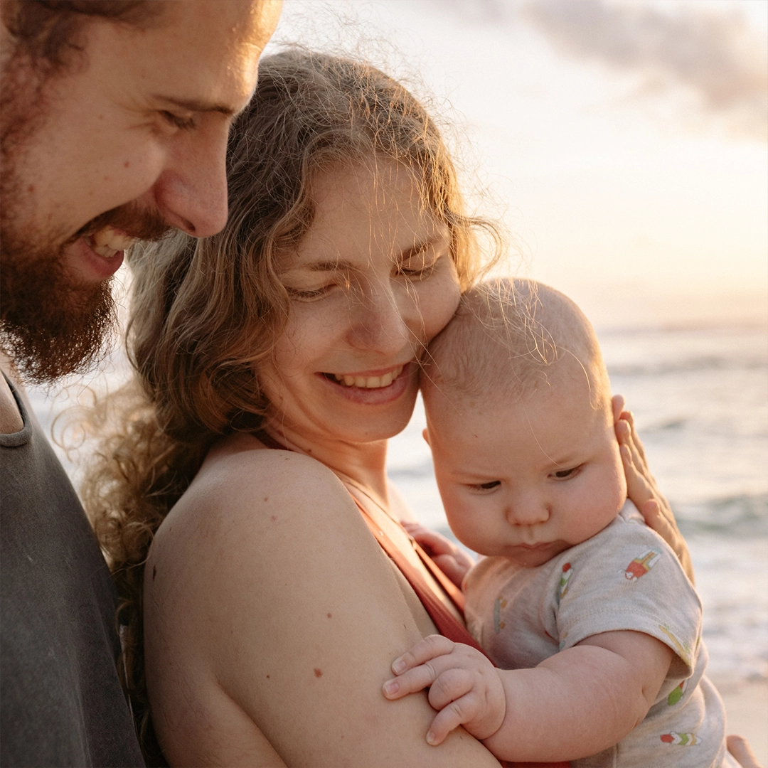 Eltern stehen am Meer und halten liebevoll ihr Baby im Arm, während sie gemeinsam einen ruhigen Moment im warmen Sonnenlicht genießen.