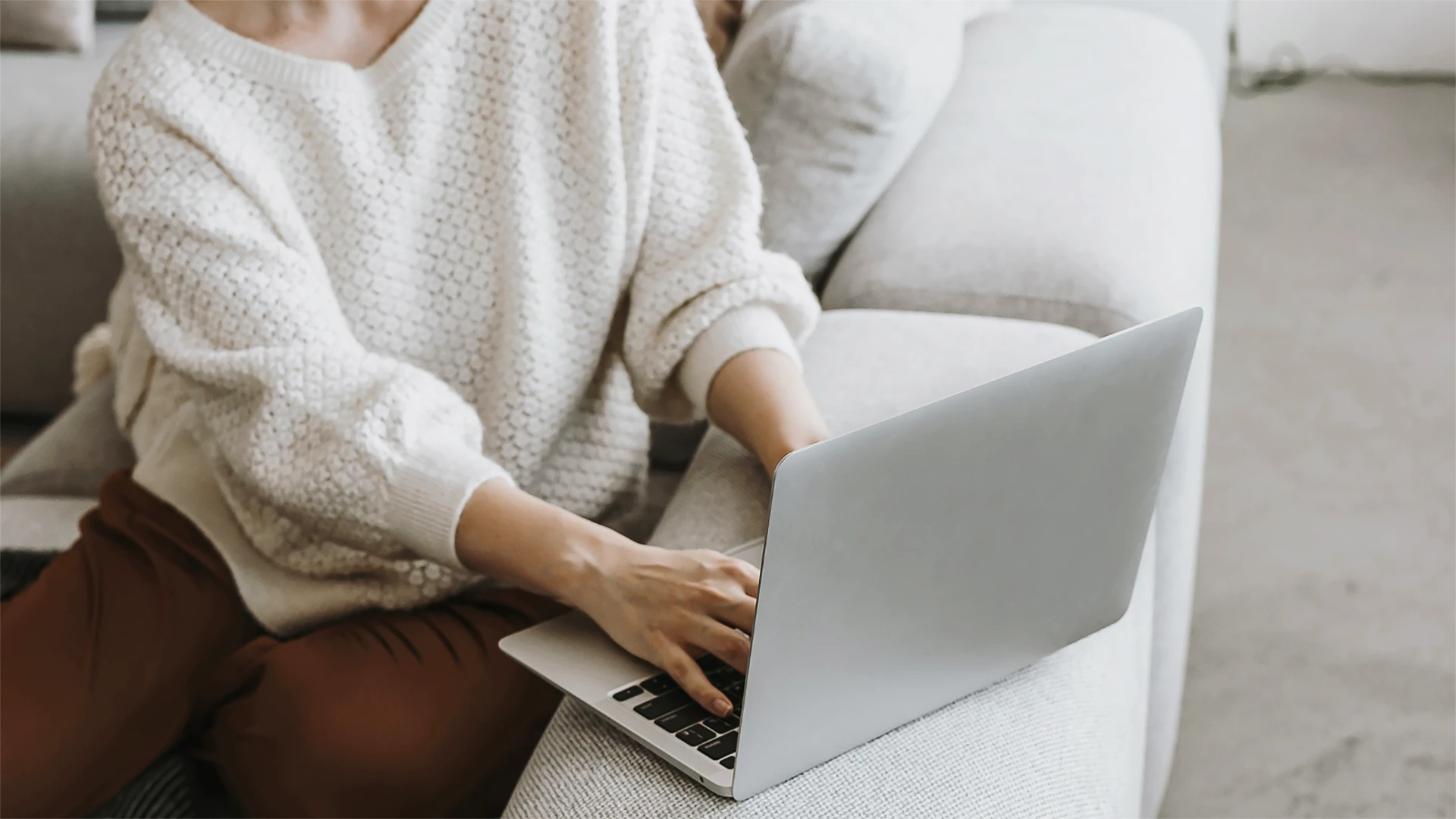 Person sitzt entspannt auf einem Sofa und arbeitet mit einem Laptop in gemütlicher Wohnatmosphäre.