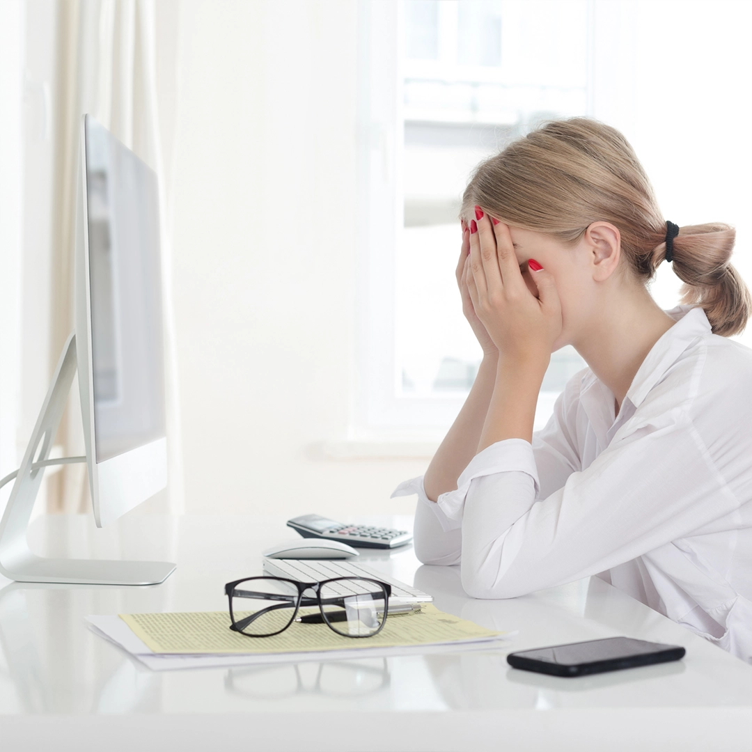 Frau sitzt gestresst am Schreibtisch vor einem Computer und hält erschöpft die Hände vor das Gesicht. Auf dem Tisch liegen Brille, Smartphone, Tastatur und Unterlagen.