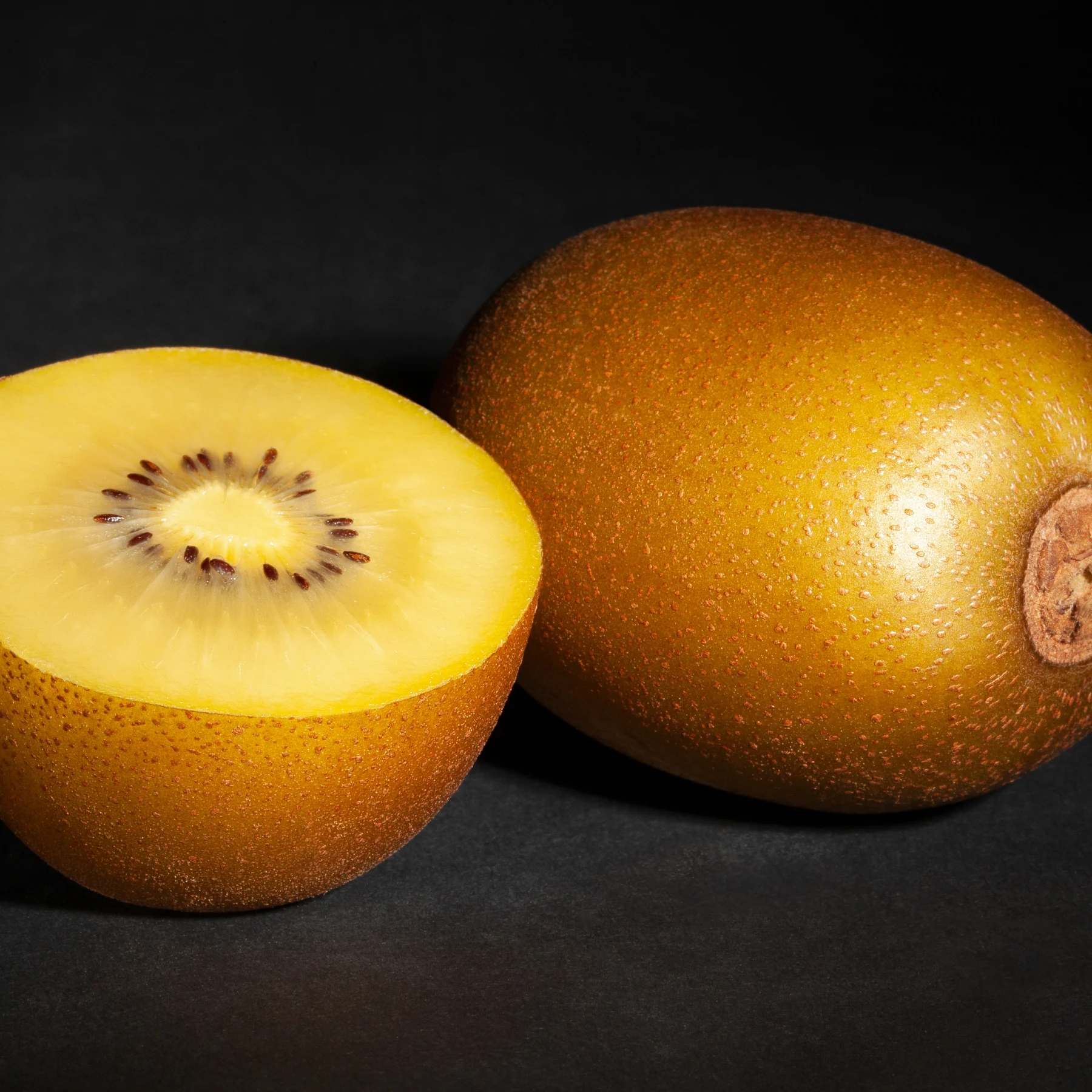 Ganze und halbierte Goldene Kiwi mit gelbem Fruchtfleisch vor dunklem Hintergrund.